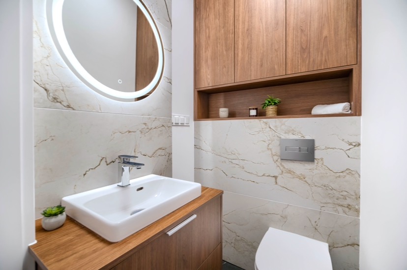 Nowoczesna łazienka z marmurowymi płytkami, okrągłym lustrem z podświetleniem LED, drewnianą szafką pod umywalką i zabudową meblową nad toaletą.