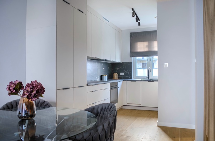 Elegancka kuchnia z białymi szafkami, czarnym marmurem na ścianie, drewnianą podłogą i szklanym stołem z fioletowymi kwiatami w wazonie.