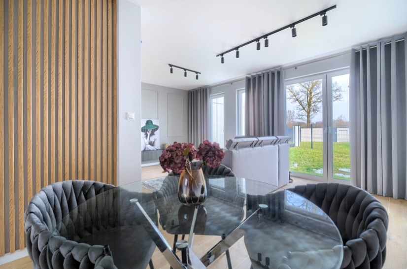 Nowoczesne wnętrze jadalni z okrągłym, szklanym stołem na metalowej podstawie, otoczonym grafitowymi krzesłami z welurową tapicerką, drewnianą ścianką działową z listew oraz oświetleniem szynowym...