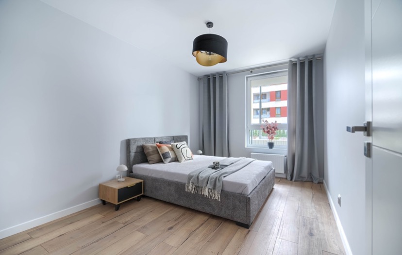 Nowoczesna sypialnia z szarym łóżkiem tapicerowanym, drewnianą podłogą i czarno-złotą lampą wiszącą, widok na budynek zza okna.