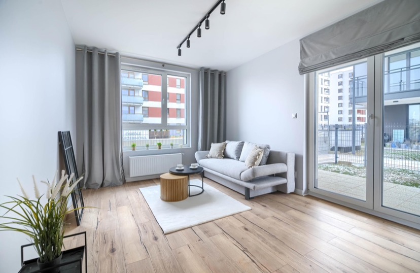 Jasne wnętrze salonu z drewnianą podłogą, szarą sofą, okrągłymi stolikami i dużym oknem balkonowym z roletą rzymską, oświetlone reflektorami szynowymi.