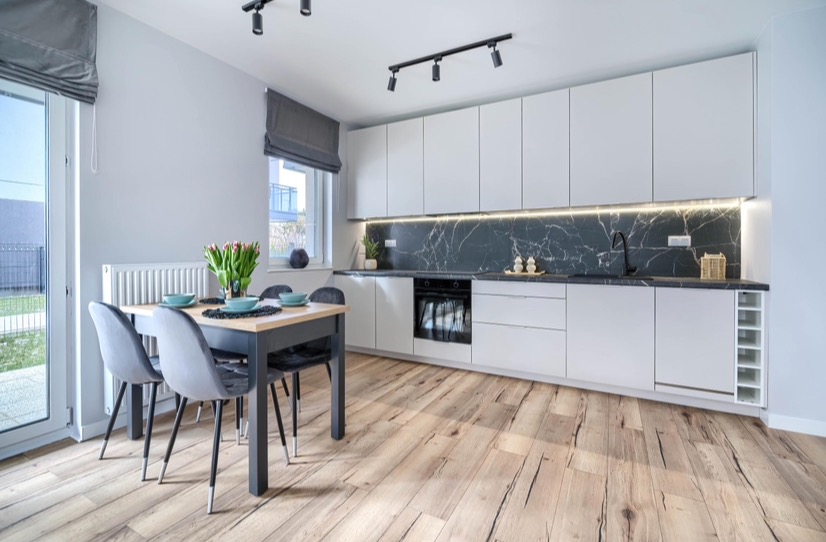 Nowoczesna kuchnia z białymi szafkami, czarnym blatem i marmurowym backsplash, oświetlenie punktowe, drewniana podłoga i stół z tulipanami.