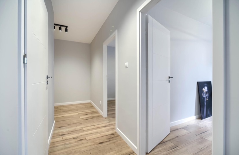 Jasne wnętrze korytarza z drewnopodobną podłogą, białymi drzwiami z poziomymi frezowaniami i szarymi ścianami, oświetlone czarnymi reflektorami sufitowymi.
