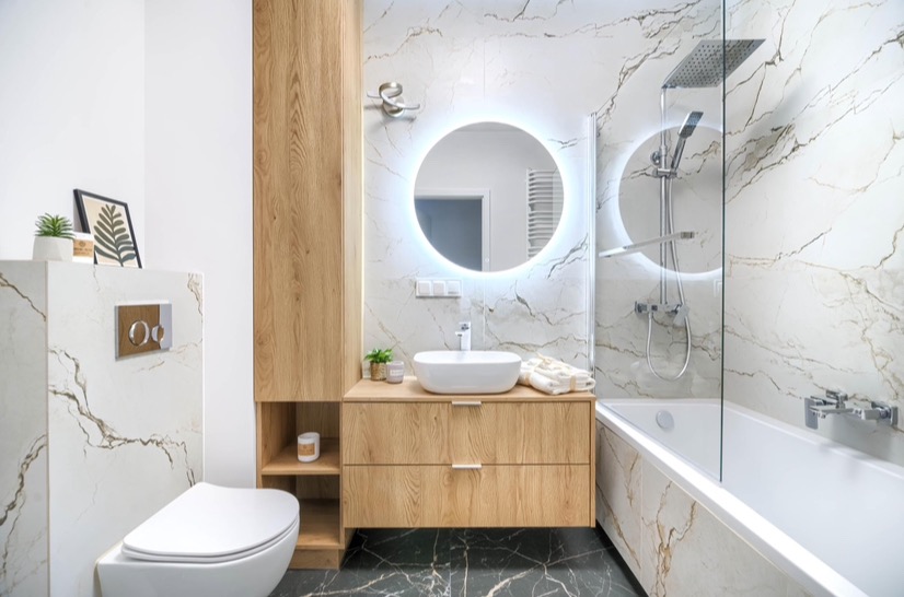 Nowoczesna łazienka z marmurową okładziną ścian i podłogą z ciemnego marmuru, okrągłe lustro z podświetleniem LED, toaleta wisząca z drewnianym panelem sterującym i zabudowa z drewnianymi szafkami.