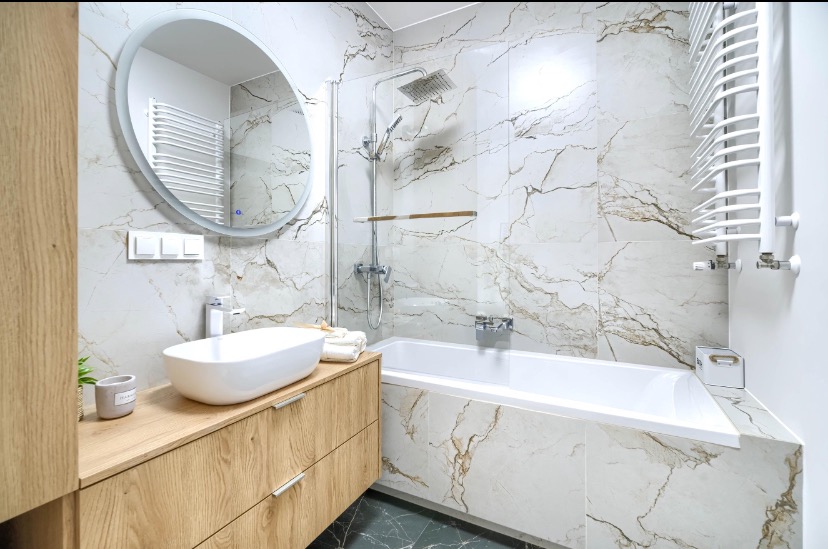 Nowoczesna łazienka z owalnym lustrem z podświetleniem LED, umywalką nablatową na drewnianej szafce, wanną obudowaną płytkami imitującymi marmur i grzejnikiem drabinkowym.