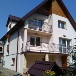 Dwupiętrowy dom z jasną elewacją, balkonami z ozdobnymi balustradami i brązowym dachem, widoczny w słoneczny dzień z fragmentem budowy w tle.