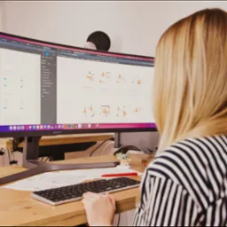 Kobieta w bluzce w paski pracuje przy zakrzywionym monitorze Samsung, na którym widoczny jest interfejs projektowania stron internetowych z elementami graficznymi. Na biurku leżą notatki...