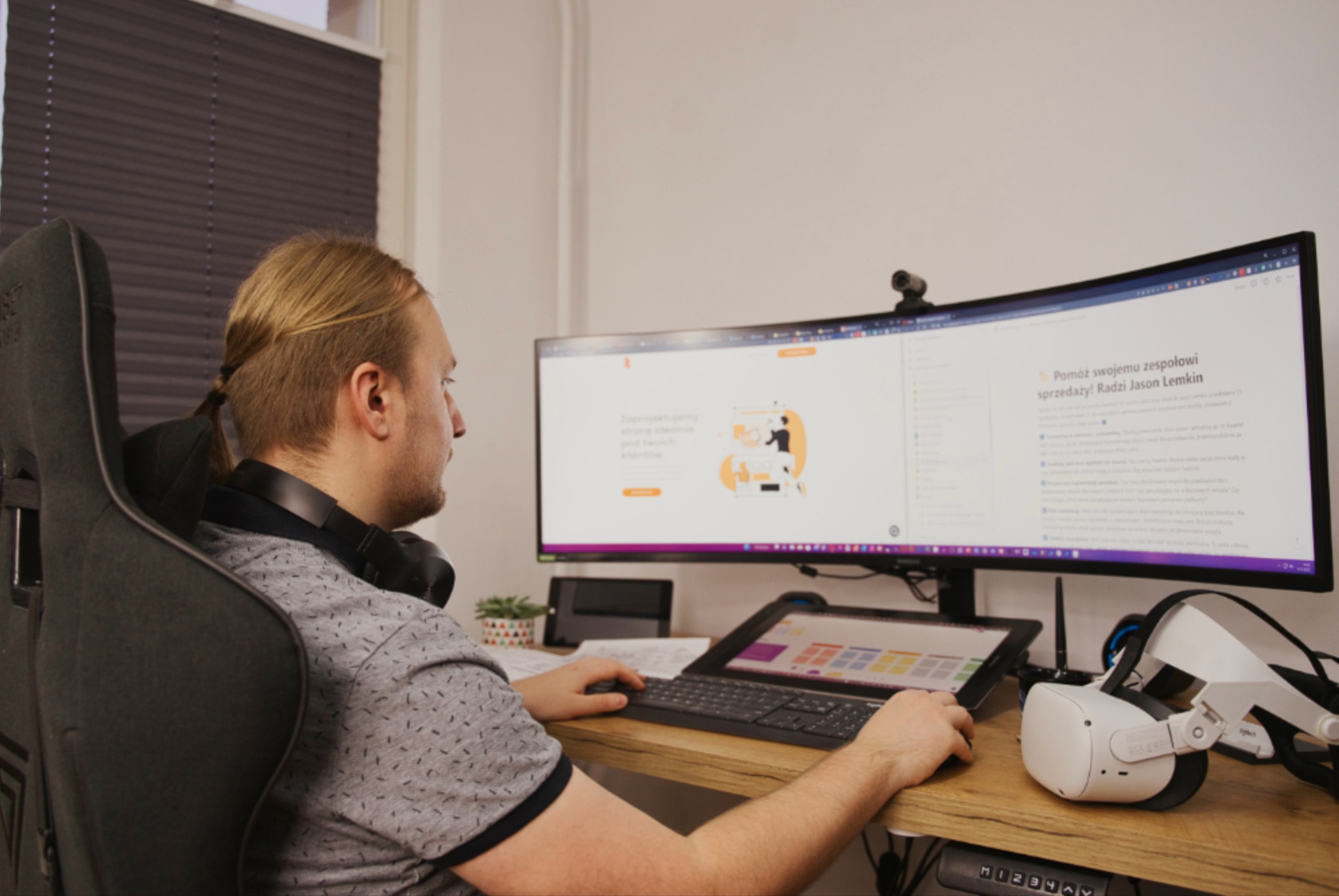 Mężczyzna z blond włosami spiętymi w kitkę, siedzi przy biurku i pracuje na dwóch monitorach, na których widoczna jest strona internetowa. Obok leżą słuchawki i gogle VR.
