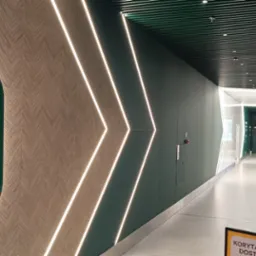Nowoczesny korytarz z podświetlanymi liniami LED w kształcie strzałek na zielonej ścianie, drewnopodobna okładzina ściany z owalnymi, zielonymi dekoracjami, sufit z metalowych listew, jasna podłoga.