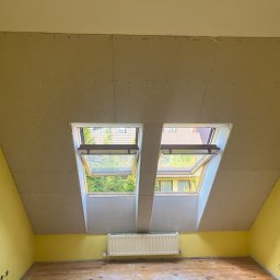 Jagi Kacper Jaglarz - Poddasze z żółtymi ścianami, gdzie na skosie dachu zamontowano płyty kartonowo-gipsowe z dwoma oknami dachowymi, przez które widać zieleń drzew i sąsiedni dom, a pod oknami znajduje się biały...