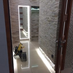 Jagi Kacper Jaglarz - Korytarz z dekoracyjną ścianą z kamienia, podświetlonym lustrem i listwą przypodłogową LED, widoczny sprzęt oświetleniowy na podłodze.