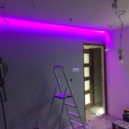 Jagi Kacper Jaglarz - Częstochowa: Wnętrze w trakcie prac wykończeniowych, widoczne szare ściany, drabina, narzędzia, prowizoryczne oświetlenie i zamontowana listwa LED z fioletowym światłem, sugerująca etap...