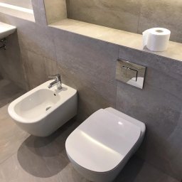 Jagi Kacper Jaglarz - Nowoczesna łazienka z szarymi płytkami imitującymi kamień, wisząca toaleta i bidet z chromowaną armaturą, rolka papieru toaletowego na półce, widoczny fragment umywalki i skrzynka narzędziowa.