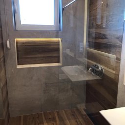 Jagi Kacper Jaglarz - Nowoczesna łazienka po remoncie: szare płytki imitujące kamień, drewnopodobna podłoga i akcenty z drewna na ścianie, przeszklona kabina prysznicowa z chromowaną armaturą, okno z roletą i wnęka...