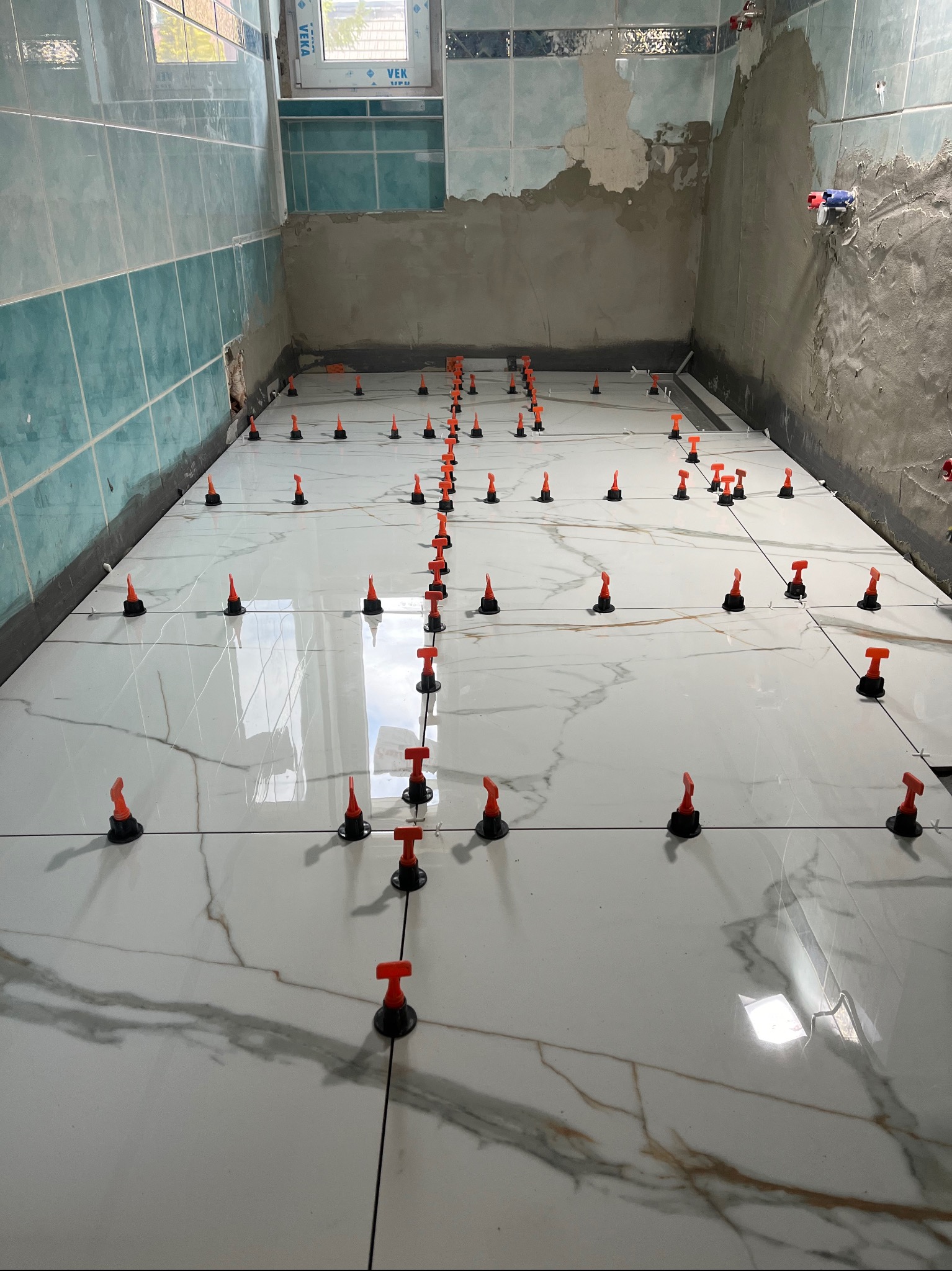 Błyszczące, białe płytki podłogowe z szarym marmurkowym wzorem, ułożone w łazience w trakcie prac wykończeniowych, z widocznymi systemami poziomowania płytek w kolorze czarno-pomarańczowym. Ściany...