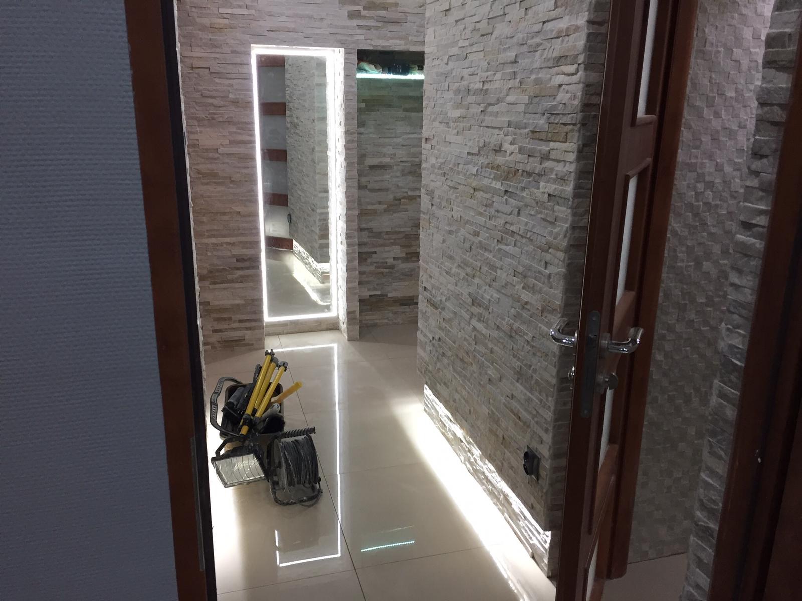 Korytarz z dekoracyjną ścianą z kamienia, podświetlonym lustrem i listwą przypodłogową LED, widoczny sprzęt oświetleniowy na podłodze.