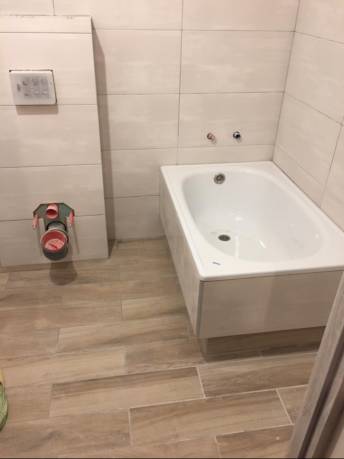 Wanna zabudowana z białymi płytkami na ścianach i drewnopodobną podłogą. Widoczne niedokończone podłączenie kanalizacyjne w ścianie.