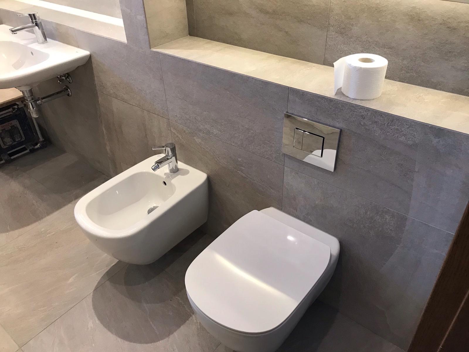 Nowoczesna łazienka z szarymi płytkami imitującymi kamień, wisząca toaleta i bidet z chromowaną armaturą, rolka papieru toaletowego na półce, widoczny fragment umywalki i skrzynka narzędziowa.