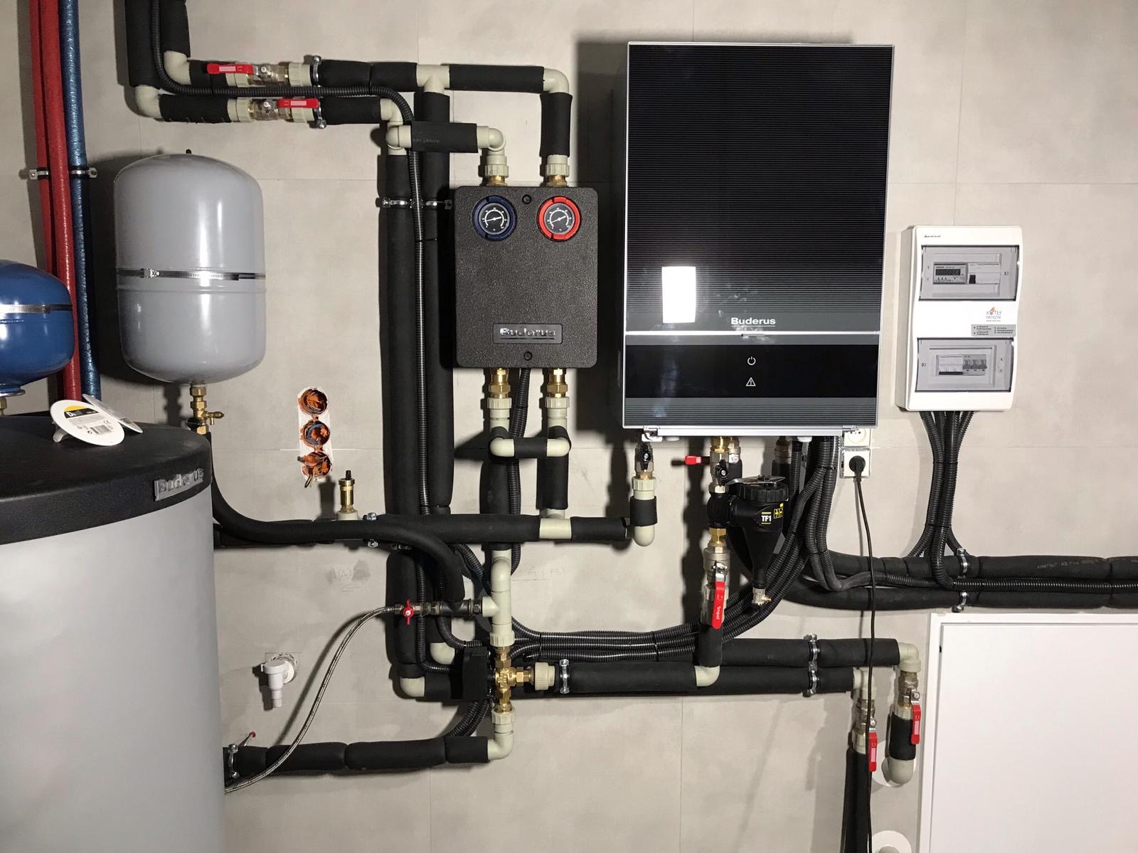 Instalacja kotła grzewczego Buderus z osprzętem, rurami w izolacji i rozdzielnicą, zamontowana na szarej ścianie z płytek ceramicznych.