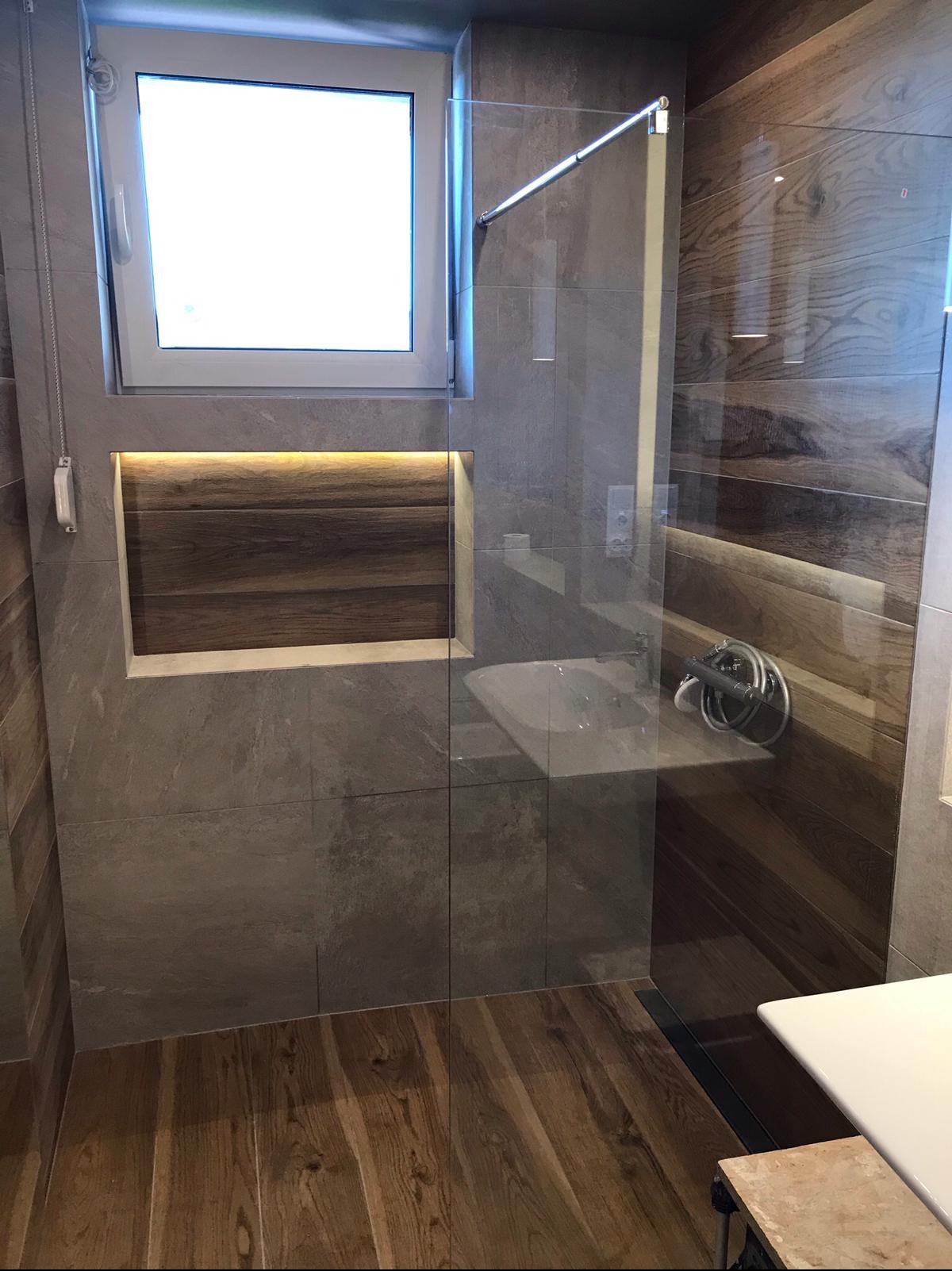 Nowoczesna łazienka po remoncie: szare płytki imitujące kamień, drewnopodobna podłoga i akcenty z drewna na ścianie, przeszklona kabina prysznicowa z chromowaną armaturą, okno z roletą i wnęka...