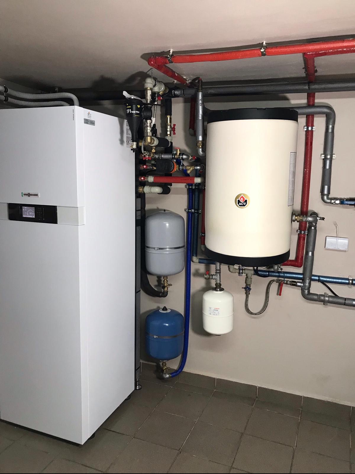 Biała pompa ciepła Viessmann, zasobnik ciepłej wody ACV i zbiorniki wyrównawcze w kotłowni z szarą podłogą z płytek i widocznymi instalacjami hydraulicznymi.