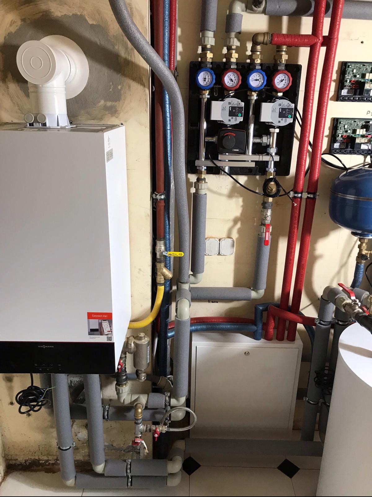 Biały piec grzewczy Viessmann z podłączonymi rurami w izolacji, manometry, pompy obiegowe i zawory na tle ściany w trakcie prac instalacyjnych.