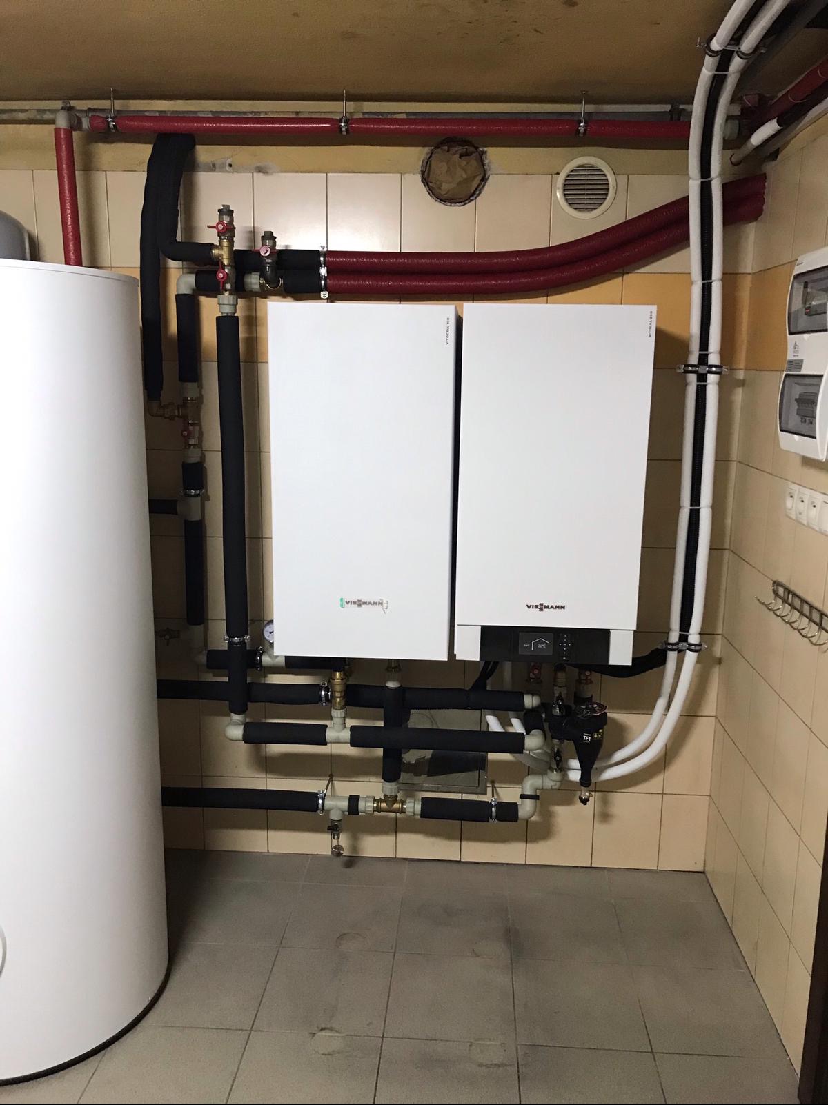 Dwa białe piece grzewcze marki Viessmann zamontowane na ścianie z beżowych płytek, z licznymi rurami w izolacji, widoczne także rury grzewcze pod sufitem i rozdzielnia elektryczna.
