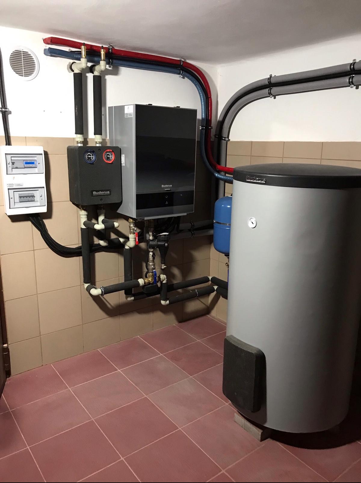 Zainstalowany, nowoczesny piec gazowy Buderus z zasobnikiem ciepłej wody, widoczne rury instalacyjne w kolorze czerwonym i niebieskim, oraz skrzynka elektryczna na ścianie wyłożonej beżowymi płytkami.