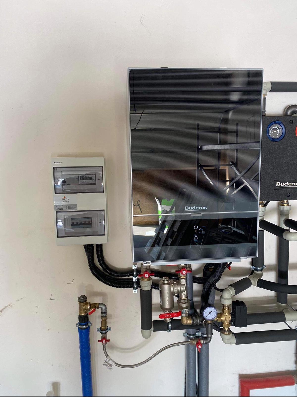 Nowoczesny piec grzewczy Buderus z czarnym, lustrzanym panelem, zamontowany na białej ścianie, z widoczną instalacją hydrauliczną i elektryczną oraz skrzynką bezpiecznikową obok.