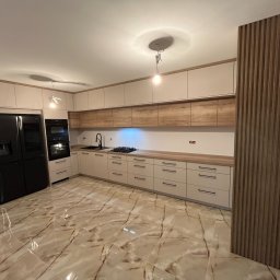 ProCraft Wojciech Pomirko - Jasna kuchnia z frontami w kolorze kremowym i drewnianym blatem, czarny piekarnik i lodówka, marmurowe płytki na podłodze, drewniana dekoracyjna ściana z pionowych listew.