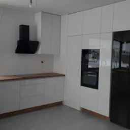 Nowoczesna, jasna kuchnia z białymi, błyszczącymi frontami szafek, drewnianym blatem i czarnymi akcentami: okap, zlewozmywak i lodówka. Za oknem zimowy krajobraz.