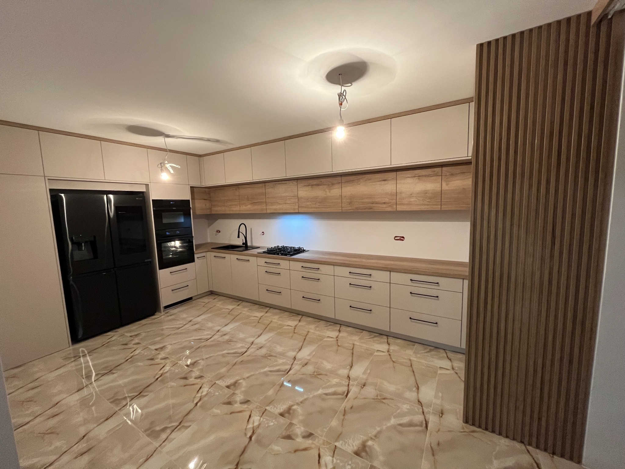 Jasna kuchnia z frontami w kolorze kremowym i drewnianym blatem, czarny piekarnik i lodówka, marmurowe płytki na podłodze, drewniana dekoracyjna ściana z pionowych listew.