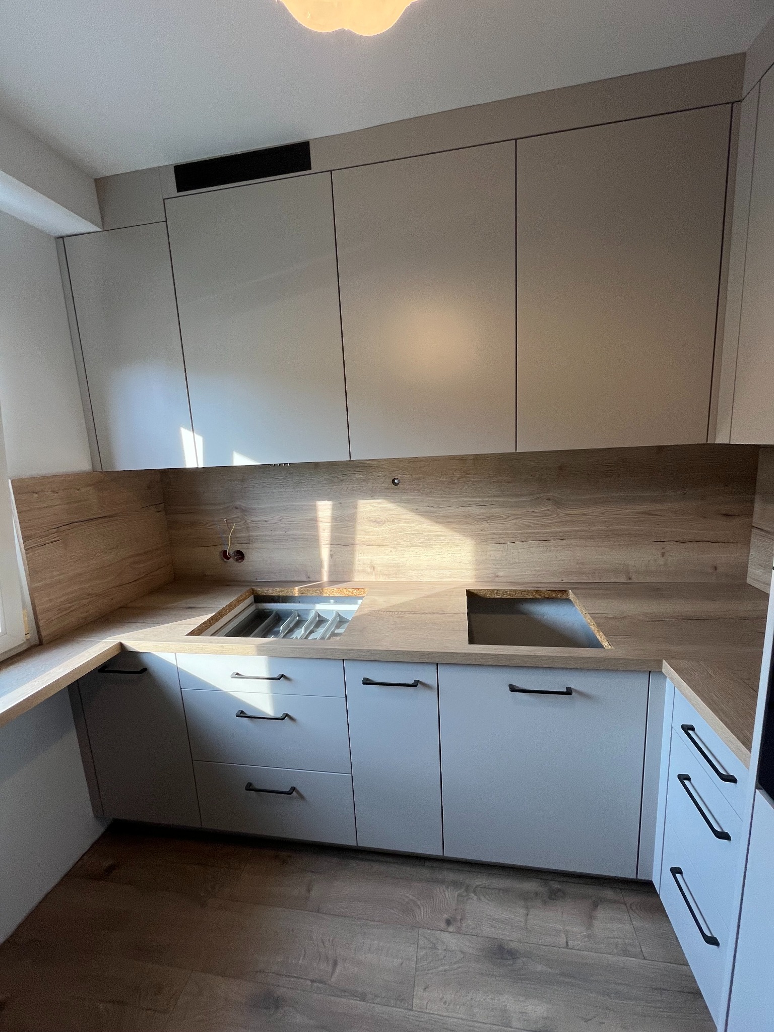 Kuchenna zabudowa na wymiar z jasnymi frontami i blatem imitującym drewno, widoczne otwory na zlewozmywak i płytę grzewczą, brak baterii i płyty.
