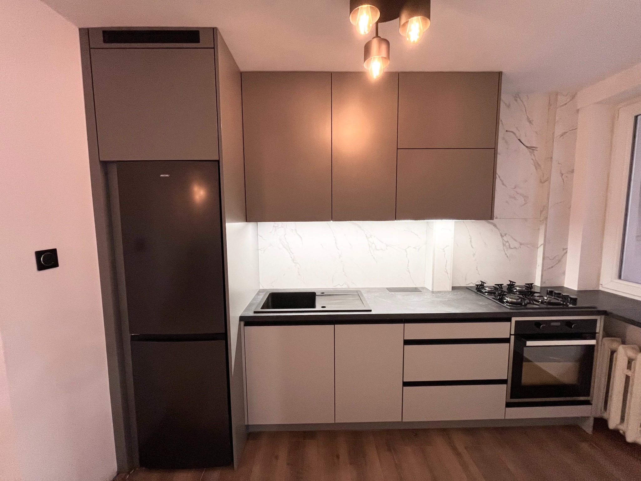 Nowoczesna kuchnia z szarymi szafkami górnymi i białymi dolnymi, czarnym blatem, marmurową ścianą nad blatem oraz wbudowanym sprzętem AGD: lodówką i piekarnikiem.