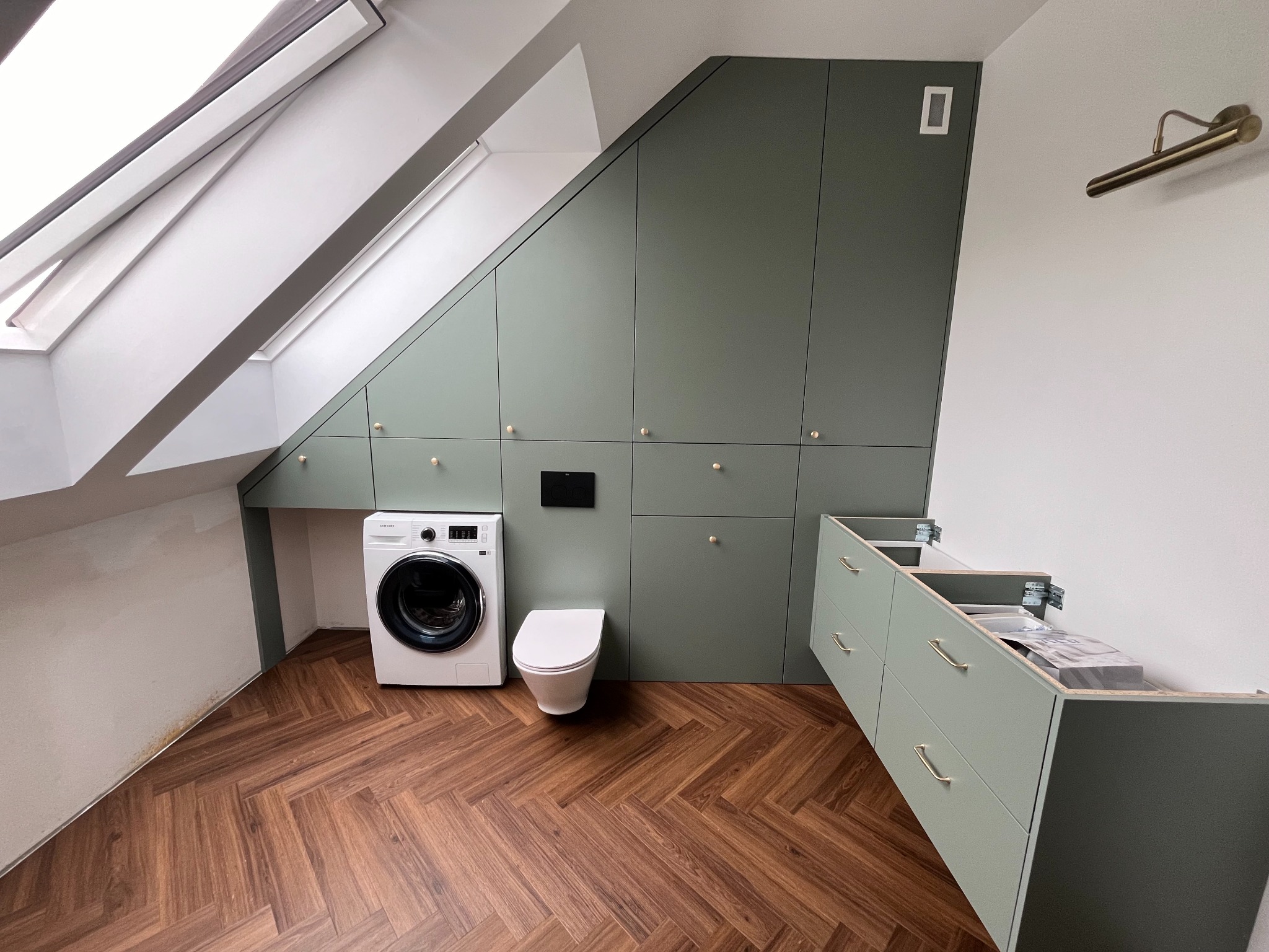 Praktyczne zagospodarowanie przestrzeni na poddaszu w Dobroszycach: zabudowa z szaf w odcieniach zieleni, dopasowana do skosów, z ukrytym WC, pralką i elementami szafki pod umywalkę, brązowa...