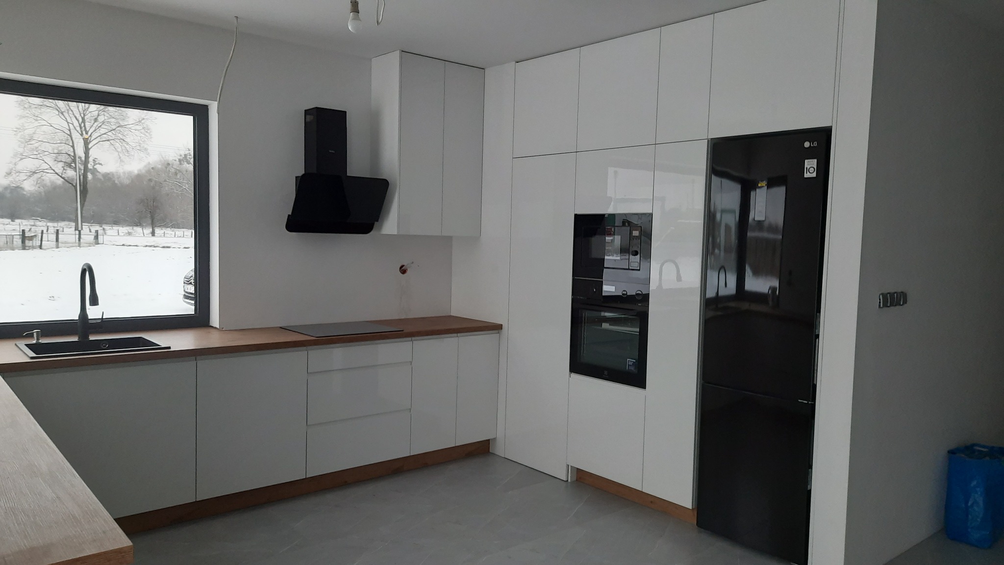 Nowoczesna, jasna kuchnia z białymi, błyszczącymi frontami szafek, drewnianym blatem i czarnymi akcentami: okap, zlewozmywak i lodówka. Za oknem zimowy krajobraz.
