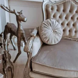 Elegancki fotel w odcieniach szarości z pikowanym oparciem i okrągłą poduszką, ozdobiony srebrnymi detalami, obok dekoracyjne figurki jeleni z brązu.