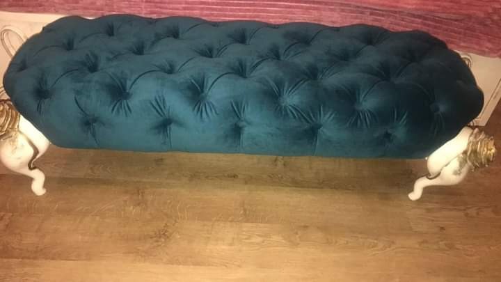 Turkusowa pufa z weluru, pikowana, na białych, rzeźbionych nogach z elementami złota, ustawiona na parkiecie.