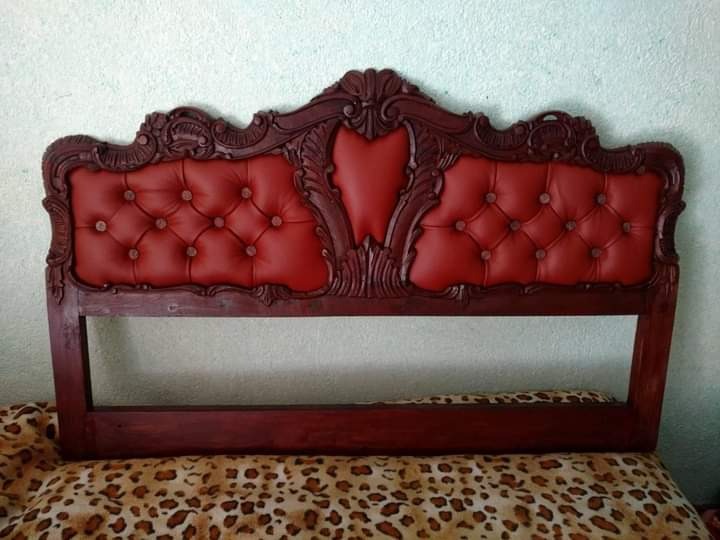 Czerwone, pikowane wezgłowie łóżka z ciemnobrązową, rzeźbioną ramą, leżące na wzorzystej tkaninie.