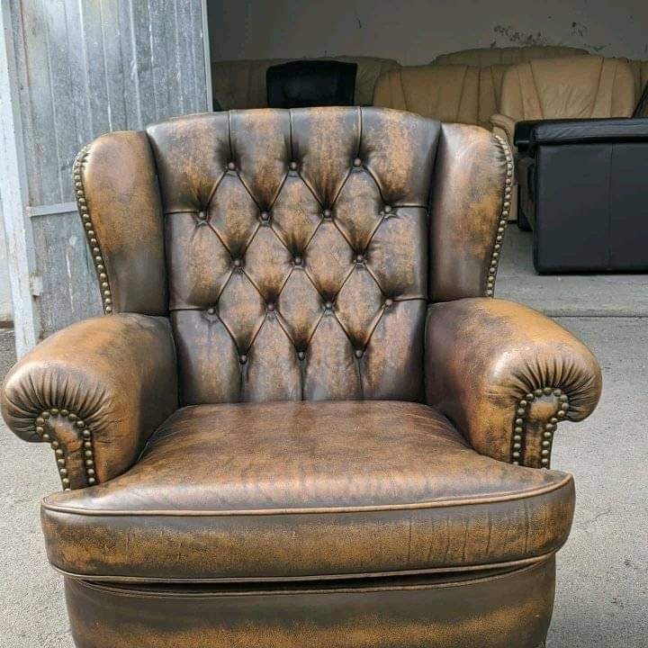 Antyczny fotel klubowy z brązowej skóry, pikowany na oparciu, zdobiony ćwiekami, z widocznymi śladami użytkowania i patyną, na tle innych mebli.