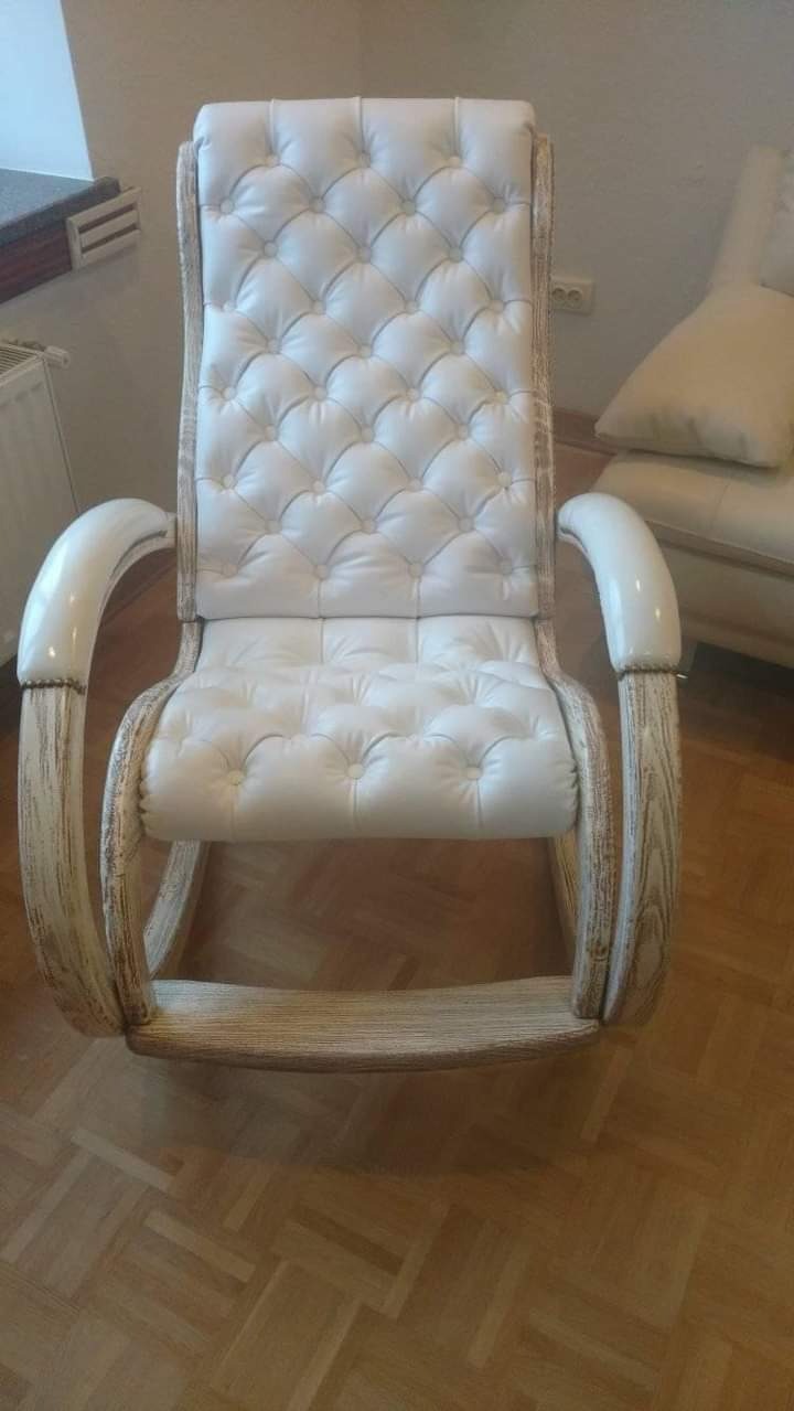 Biały, skórzany fotel bujany z pikowanym oparciem i podłokietnikami, na drewnianej ramie o przecieranym wykończeniu, stojący na parkiecie.