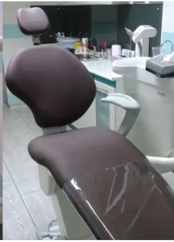 Brązowy fotel dentystyczny z przezroczystą folią ochronną na siedzisku, widoczny w gabinecie stomatologicznym z białymi meblami i umywalką.