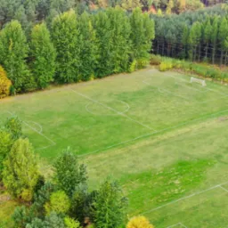 Widok z lotu ptaka na zielone boisko do piłki nożnej otoczone lasem, z widocznymi bramkami i liniami wyznaczającymi pole gry.
