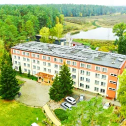 Widok z lotu ptaka na trzypiętrowy pensjonat z panelami słonecznymi na dachu, otoczony zielonymi drzewami i niewielkim jeziorem w tle.