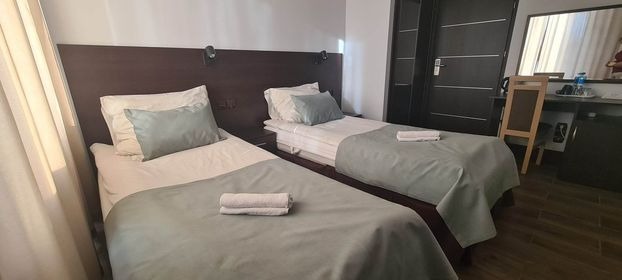 Pokój hotelowy z dwoma pojedynczymi łóżkami, szarozielona pościel, ciemnobrązowe wezgłowie, oświetlenie punktowe, widok na biurko i szafę w tle.