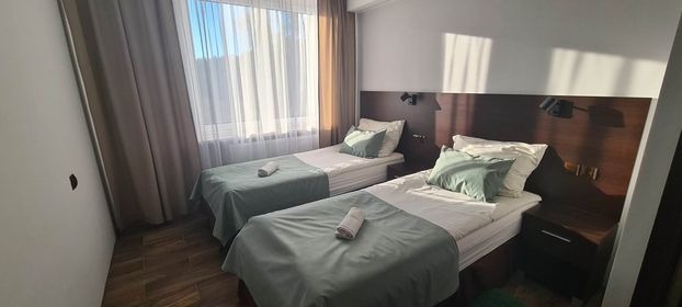 Pokój hotelowy z dwoma pojedynczymi łóżkami, zielonymi narzutami i poduszkami, drewnianym zagłówkiem i szafkami nocnymi, oświetlony naturalnym światłem wpadającym przez okno z jasnymi zasłonami.