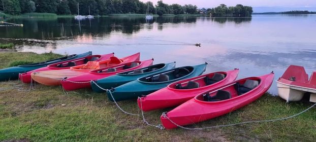 Kolorowe kajaki i rower wodny, przygotowane do wypożyczenia nad jeziorem w Szeligach, w tle widoczne żaglówki i linia brzegowa.