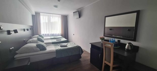 Wnętrze pokoju hotelowego z trzema pojedynczymi łóżkami, biurkiem z lustrem, czajnikiem elektrycznym i kwiatami w wazonie.