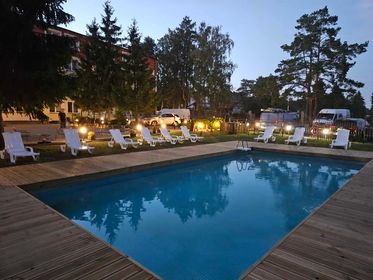 Oświetlony basen zewnętrzny z drewnianym pomostem, otoczony leżakami i drzewami o zmierzchu.