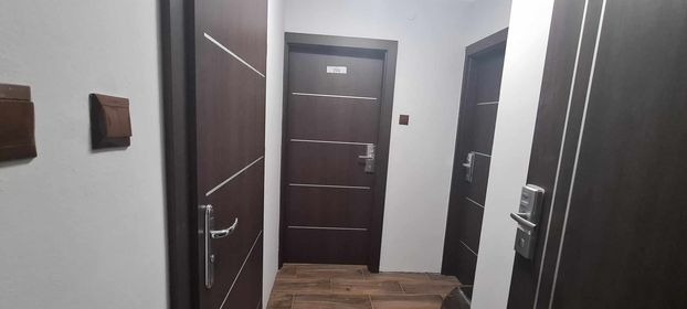 Korytarz hotelowy z trzema ciemnymi drzwiami z numerami pokoi i elektronicznymi zamkami, oświetlenie górne.